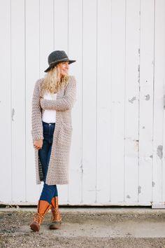 a woman standing in front of a white wall wearing a long cardigan and boots