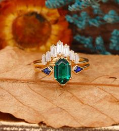 an emerald and diamond ring sitting on top of a brown leaf in front of flowers