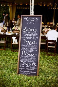 a chalkboard menu sign sitting on top of a grass covered field next to tables