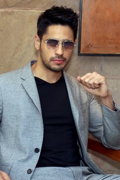 a man wearing sunglasses sitting on a bench with his hand in his pocket and looking at the camera