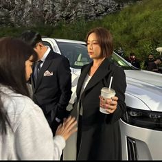 a woman standing next to a man in front of a white car holding a drink