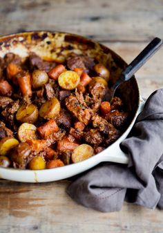 a white bowl filled with meat and potatoes on top of a wooden table next to a gray napkin