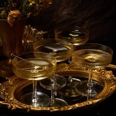 four champagne glasses sitting on a tray with gold trimmings and flowers in the background