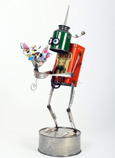 a tin can robot holding a flower in it's hand and standing on top of a metal base