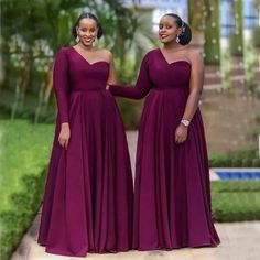 two women in long purple dresses standing next to each other on a sidewalk with trees and bushes behind them