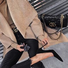 a woman sitting on a bench with her legs crossed and wearing black jeans, a beige coat and heels