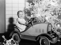 black and white photograph of a baby in a toy car next to a christmas tree