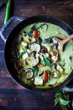 a pan filled with green soup and vegetables on top of a wooden table next to a spoon