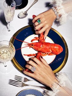 two hands holding a lobster on a plate with silverware and wine glasses around it