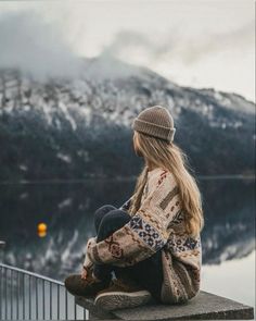 a woman sitting on top of a stone wall next to a body of water with mountains in the background