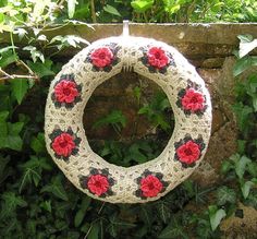 a knitted wreath with red flowers hanging on a stone wall surrounded by greenery