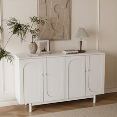 a white sideboard with two plants on top and a lamp in the corner next to it