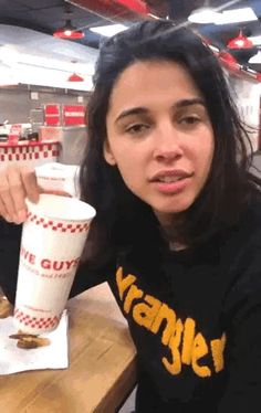 a woman sitting at a table with a paper cup in her hand and food on the counter behind her