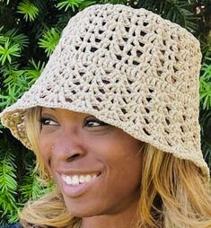 a smiling woman wearing a crocheted hat in front of some bushes and trees