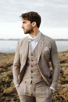 a man wearing a suit and bow tie standing in front of the ocean with his hands in his pockets