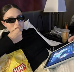 a woman laying in bed with a bag of chips on her lap and looking at the screen