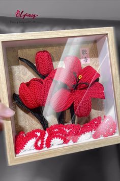 a person holding up a crocheted red flower in a frame with the word love written on it