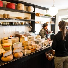 two women standing in front of a counter filled with cheeses and other food items