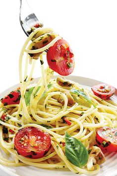 spaghetti with tomatoes and spinach being lifted by tongs from a white plate on a white table