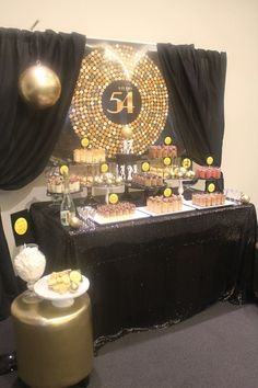 a table with desserts on it in front of black drapes and gold decorations