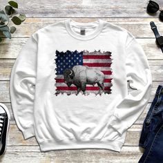 Experience the powerful combination of nature and patriotism with our outstanding Bison graphic t-shirt or sweatshirt. Our exclusive design showcases the imposing appearance of an American bison against the backdrop of the impressive American flag. This bison graphic shirt is not just a piece of clothing, it is an expression of love for wildlife and proud national pride. The high-quality sweatshirt not only offers a visually appealing display, but also unparalleled comfort for your active outdoor lifestyle. Great bison graphic shirt or sweatshirt for bison lovers, nature lovers, America lovers, hunters, camping lovers, bison breeders, people who spend their next vacation in America or love the country of America. Great design also for American Independence Day or Patriot`s Day.  For those American Bison, American Independence, Camping Lovers, Animal Shirts, Graphic Shirt, Gift For Dad, Showcase Design, Relaxed Style, Animal Design