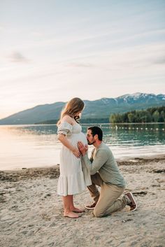 a man kneeling down next to a pregnant woman on the beach with mountains in the background