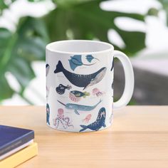 a coffee mug sitting on top of a wooden table next to a book and plant