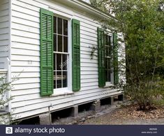 an old white house with green shutters on the front and side windows, overgrown by trees