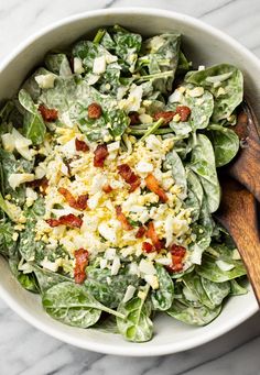 a salad with bacon and cheese in a white bowl on a marble counter top next to a wooden spoon