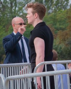 two men standing next to each other in front of a metal barricade with trees in the background