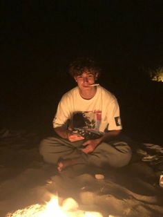 a young man sitting in front of a campfire with a book on his lap