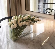 white tulips in a square glass vase on a marble table with a mirror behind it