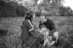 a man and woman sitting in tall grass with a dog on their lap looking at each other