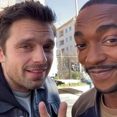 two men standing next to each other in front of a building and one is giving the peace sign