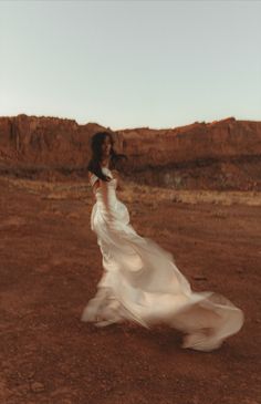 a woman in a long white dress is walking through the desert with her hair blowing in the wind