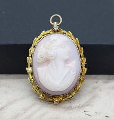 Introducing an exquisite 10k Cameo Brooch Pendant! Crafted with a light pink shell, this stunning piece features an intricately carved portrait of a young woman, boasting a roman nose and beautiful hair adorned with a delicate flower. The antique shell cameo is bezel set in 10k yellow gold fillagree with a diamond accent at the top that adds a touch of elegance. The brooch attaches with a 'C' clamp and has an unusual apparatus for a safety clasp. At the top of the cameo is a folding ring bail fo Luxury Cameo Brooch For Wedding, Carved Yellow Gold Brooches For Wedding, White Cameo Brooch For Wedding, White Cameo Brooches For Wedding, White Cameo Wedding Brooches, Cameo Pendant Brooch For Wedding, Cameo Pendant Brooches For Wedding, Elegant Pink Brooches For Collectors, Roman Nose