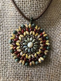 This colorful hand-beaded Mandala Necklace is made using many different style beads, and as you can see, very colorful!  It is warm colors, which include greens, browns, deep red, and brass colored beads. These include Super Duo beads, as well as assorted round beads in various sizes, and a large cut bead as the center piece. It is quite flexible, as shown in the one picture, but lays flat against your body. It measures 1-3/4 " inches in diameter, not including the beaded bale. It also includes Bohemian Brown Beaded Necklace, Bohemian Brown Beaded Round Necklaces, Bohemian Round Brown Beads, Bohemian Beaded Necklaces With Faceted Beads, Bohemian Brown Beaded Necklaces With Oval Beads, Brown Beaded Necklace With Colorful Oval Beads, Handmade Brown Czech Glass Beads, Earthy Multicolor Gemstone Beaded Necklaces, Earthy Spacer Beads For Jewelry Making