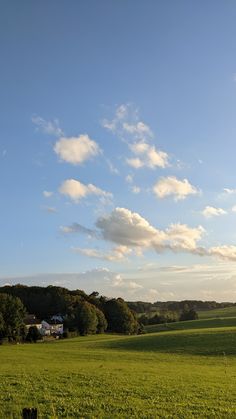 an open field with trees and houses in the distance under a blue sky filled with clouds