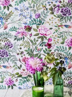 two green vases with flowers in front of a floral wallpapered background on a table