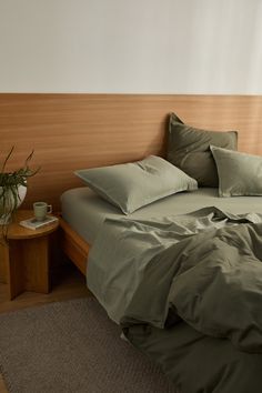 a bed with green sheets and pillows in a room next to a plant on a table