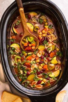 a crock pot filled with vegetable soup and some sliced bread on the table next to it