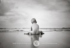 a black and white photo of a baby sitting on the beach looking out at the ocean