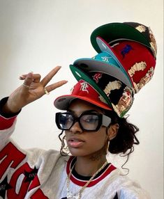 a woman wearing glasses and a hat making the peace sign with her hands while standing in front of a white wall