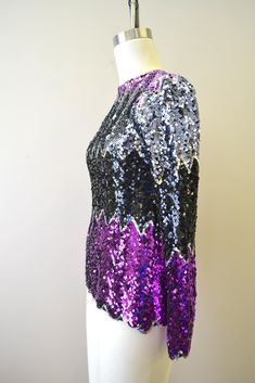 "Description: 1970s black, silver, and purple sequined shirt in wide, chevron stripes. Made on an acrylic knitted base, so there is a tiny amount of stretch to the fabric, as well as some holes from the mesh-like base. 42% acetate, 46% acrylic, 12% cotton. Long sleeves and a scalloped hem. Back button at neck. Condition: In great condition. There are no holes or stains. Designer: n/a Stated Size: Small Measurements: Armpit to armpit: 16.5\" Shoulder to shoulder: 14\" Sleeve length: 24\" Overall Purple Winter Party Top, Fitted Purple Sequin Top, Purple Tops For Evening Party Season, Purple Tops For Evening And Party Season, Purple Evening Tops For Party Season, Purple Top For Evening And Party Season, Glamorous Purple Sequin Top, Sequined Top, Sequin Shirt