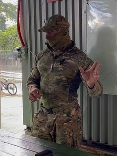 a man in camouflage standing next to a table with his hands out and wearing a mask