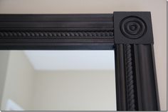 a black mirror frame with an ornate design on the bottom and sides, in front of a white wall