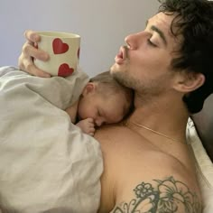 a man laying in bed holding a baby and drinking from a cup with hearts on it