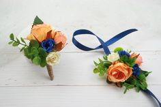 two boutonnieres with orange and blue flowers are on a white wooden surface