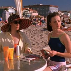 two women are sitting at an outdoor table on the beach, one reading a book