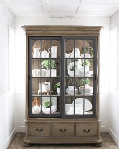 an old china cabinet with glass doors and white dishes on it's shelves in the corner